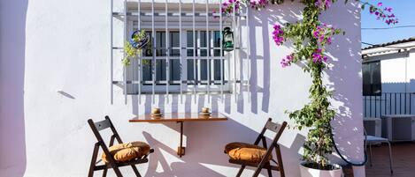 An outdoor seating area with two chairs and a table, a window with a metal grill, and a potted plant with purple flowers.