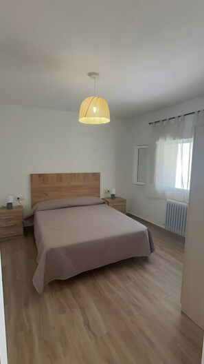 A bedroom with a wooden headboard, a bed with a beige bedspread, a bedside table with a lamp, and a window with curtains.