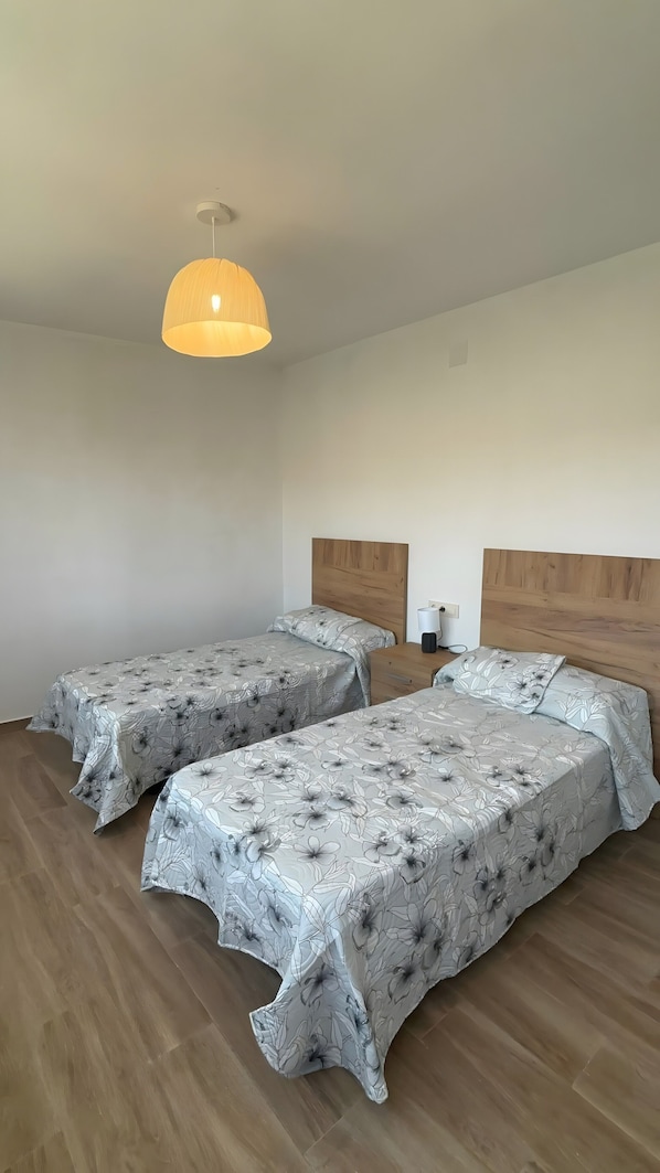 A room with two beds, a wooden headboard, a bedside table with a lamp, and a hanging light fixture.