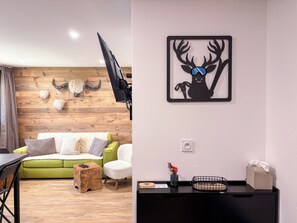 A modern living room with a wooden wall, a green sofa, a white armchair, a small wooden stool, and a black TV stand with a mounted TV. - Les Caribous - 5 min from Megève, Lake and Espace Diamant (Praz-sur-Arly)