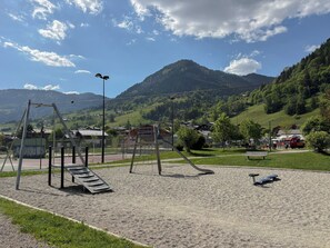 A playground with swings and a slide, surrounded by a park with benches and a mountain backdrop. - Les Caribous - 5 min from Megève, Lake and Espace Diamant (Praz-sur-Arly)