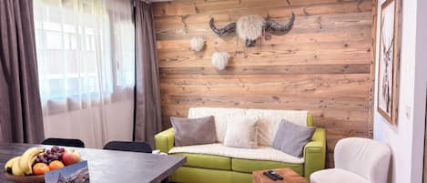 A modern living room with a wooden wall, a green sofa, a white armchair, a wooden coffee table with fruit, and a remote control on a wooden stool.