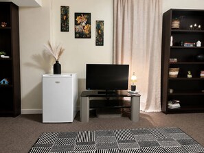 Ein Wohnzimmer mit Flachbildfernseher, einem kleinen, weißen Kühlschrank, einem Bücherregal und einem gemusterten Teppich.