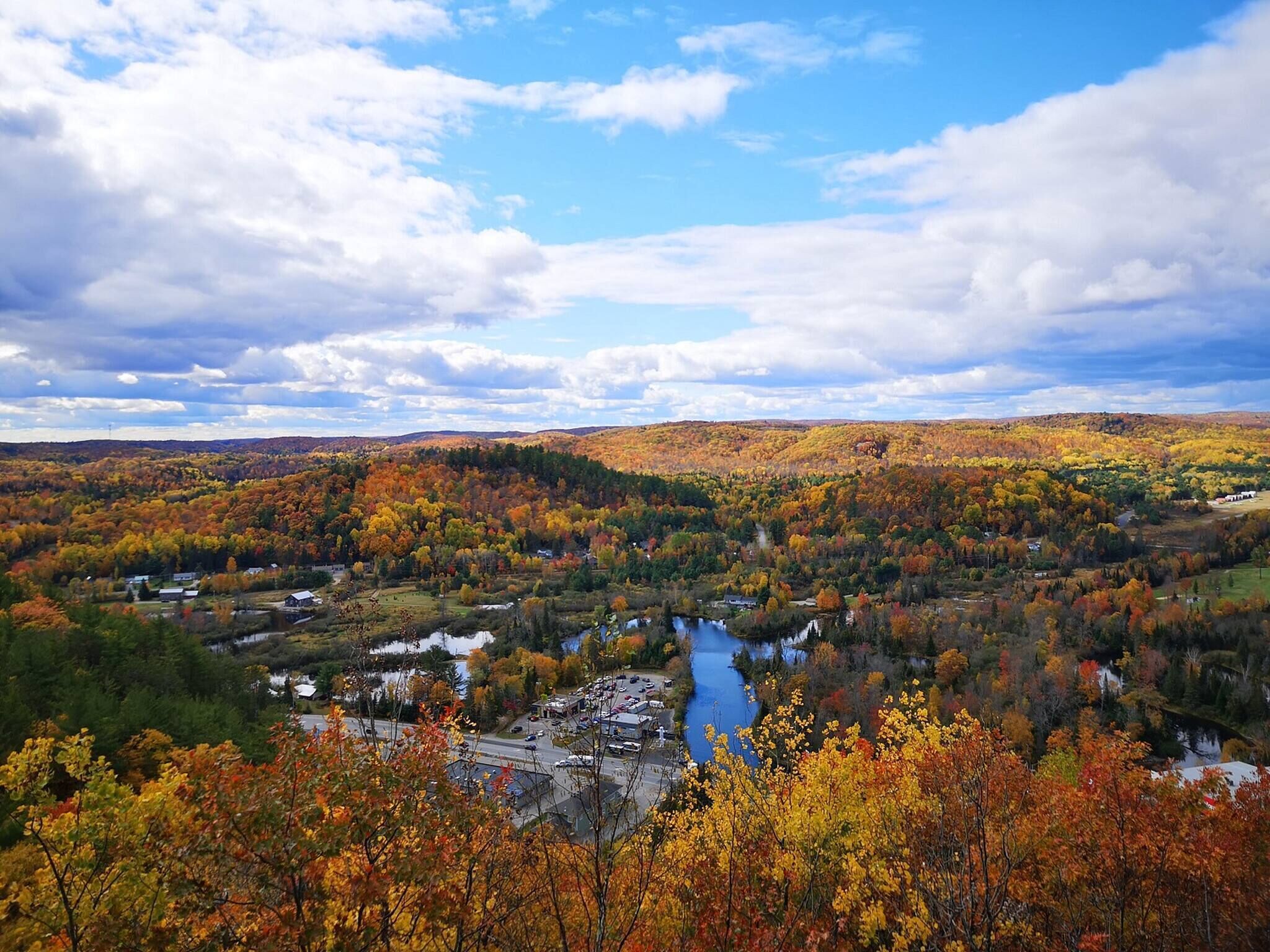 A river flowing through a valley with autumn foliage, surrounded by hills and a partly cloudy sky.