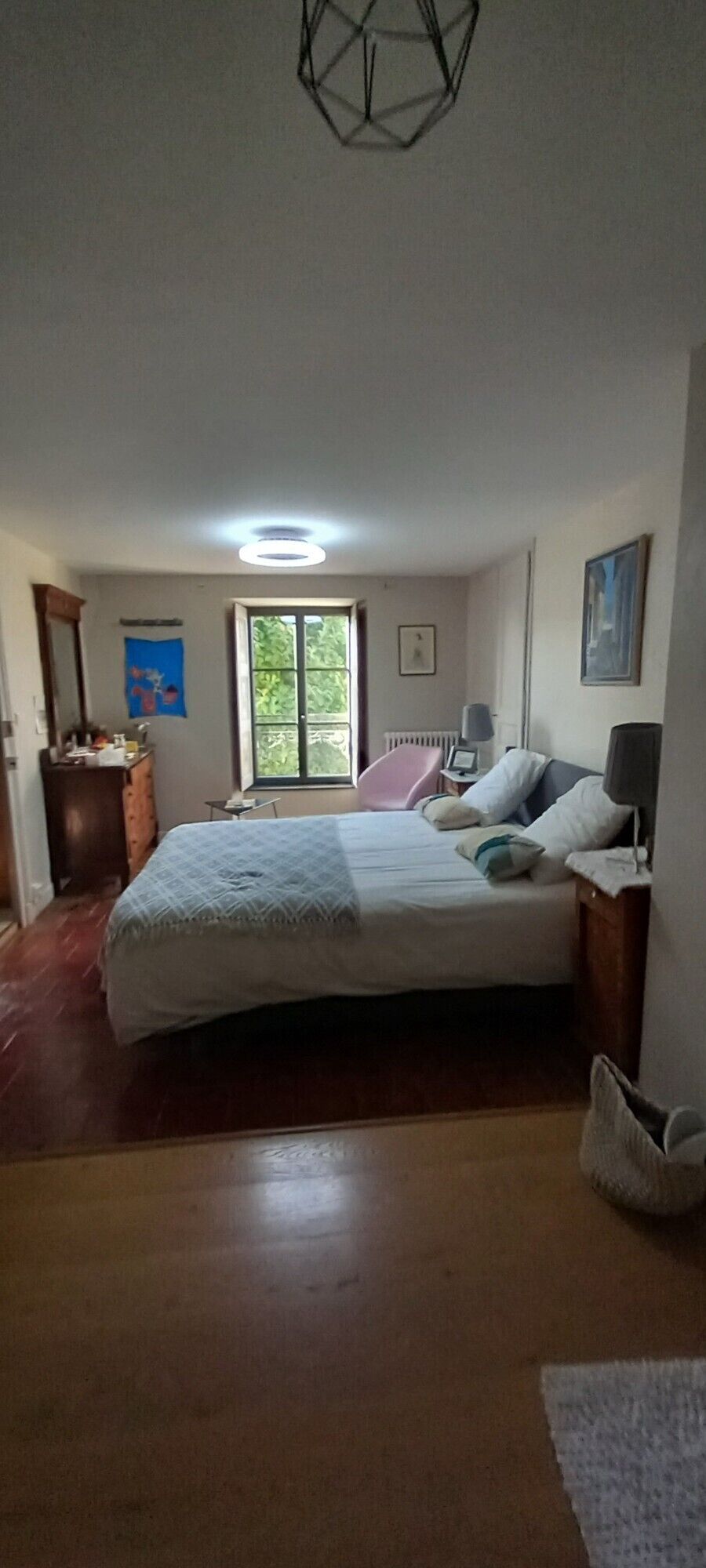 Un dormitorio con cama, mesitas de noche, escritorio, silla, radiador y ventana con vistas al verdor.