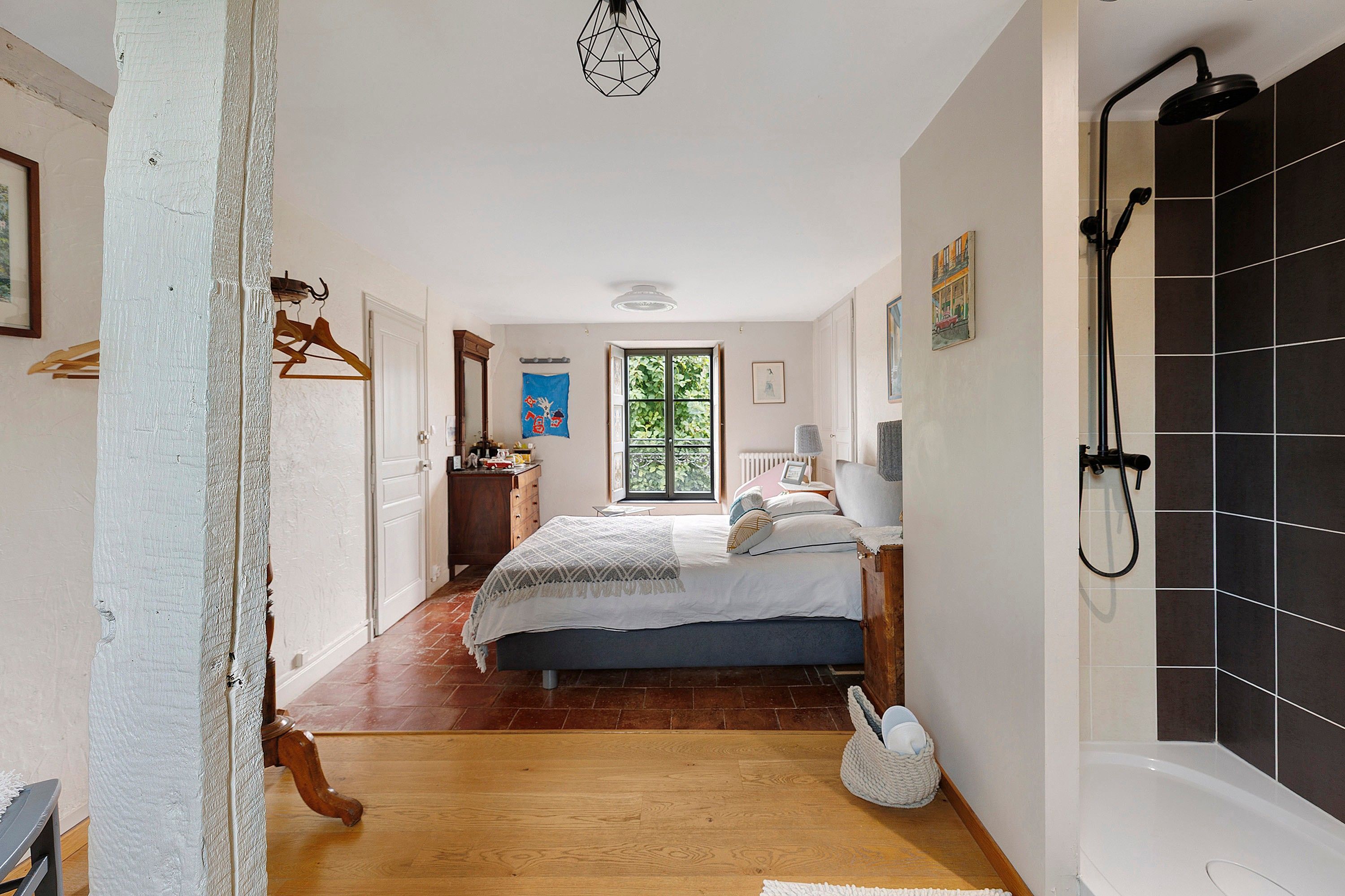 Een slaapkamer met een bed, nachtkastje, hanglamp, raam en een badkamer met douche.