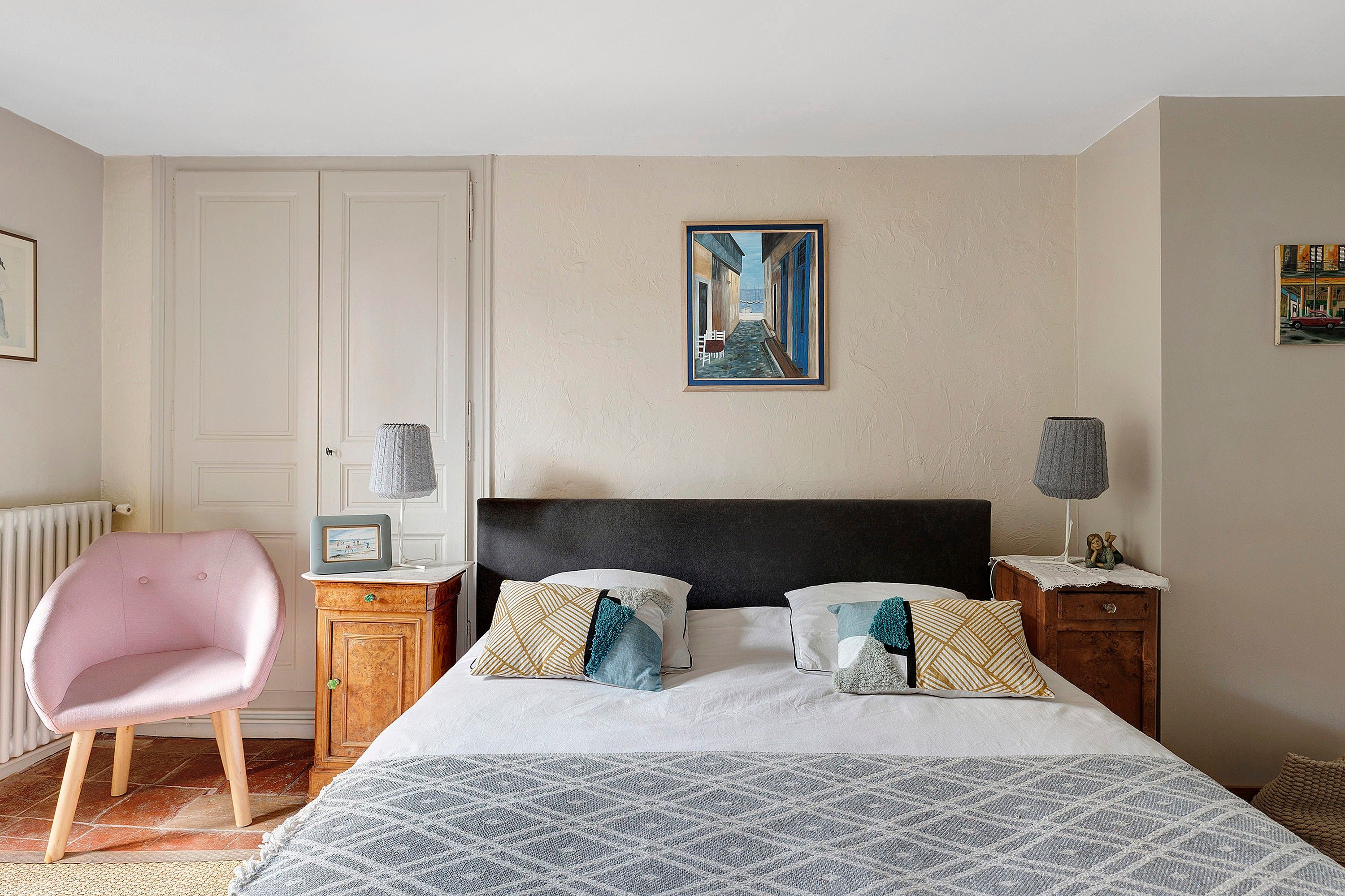 Quarto com cama, cadeira, mesa de cabeceira, abajur e um quadro na parede.