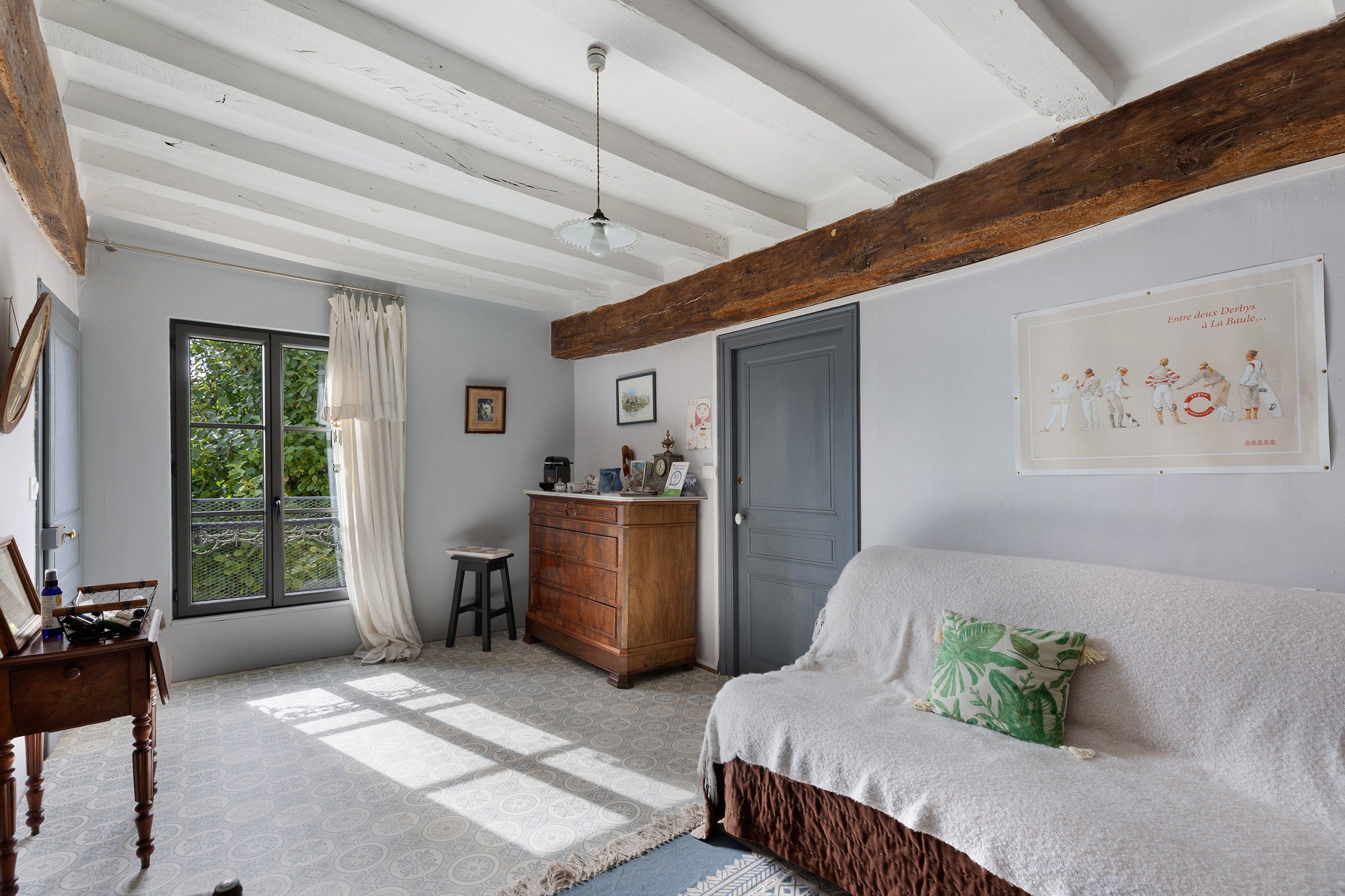 Une pièce avec un plafond en bois, une fenêtre avec des rideaux, une commode en bois, un canapé avec un coussin et un mur orné d’un tableau encadré.