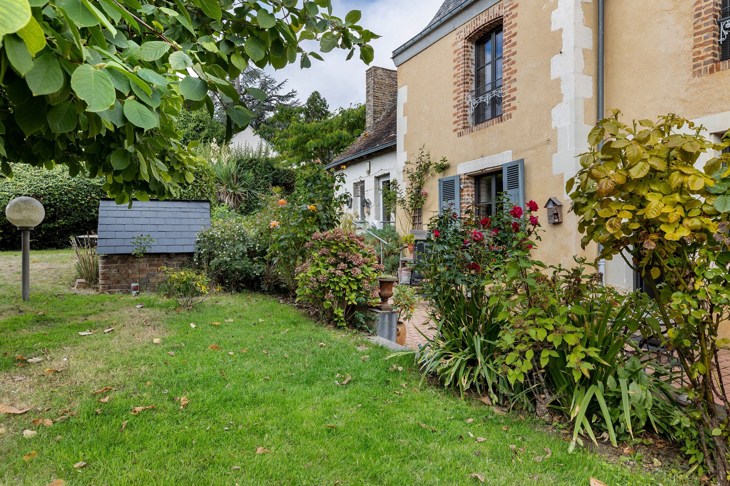 Un jardin résidentiel avec une pelouse, diverses plantes et une maison en briques dotée de volets.