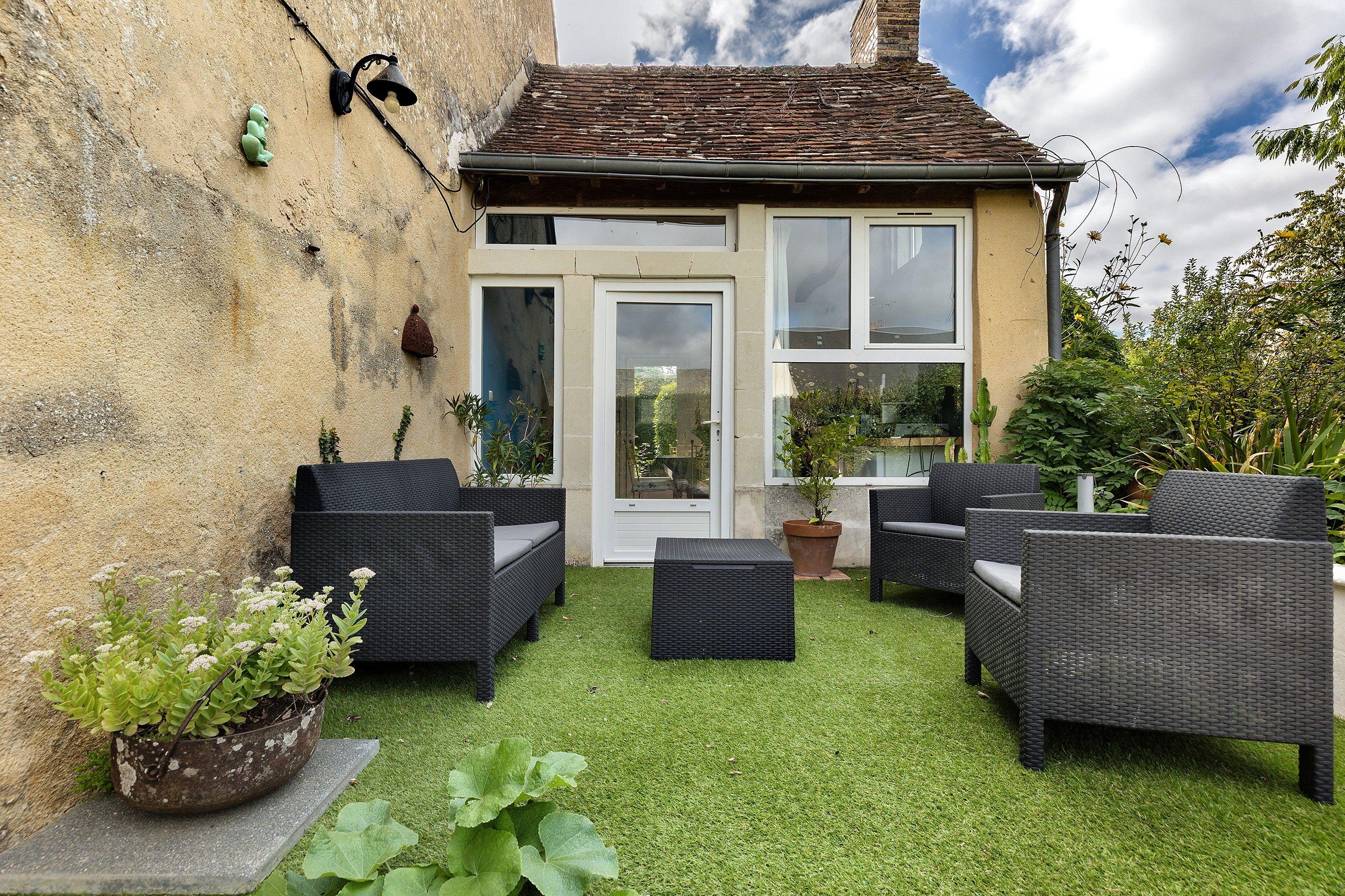 A small house with a tiled roof, a white door, and a window. There are wicker furniture pieces and a potted plant on the lawn.