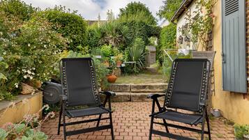 Deux chaises noires sont placées face à un jardin, avec un chemin en briques qui le traverse, et l’ensemble est entouré de différentes plantes et d’une petite structure en bois.