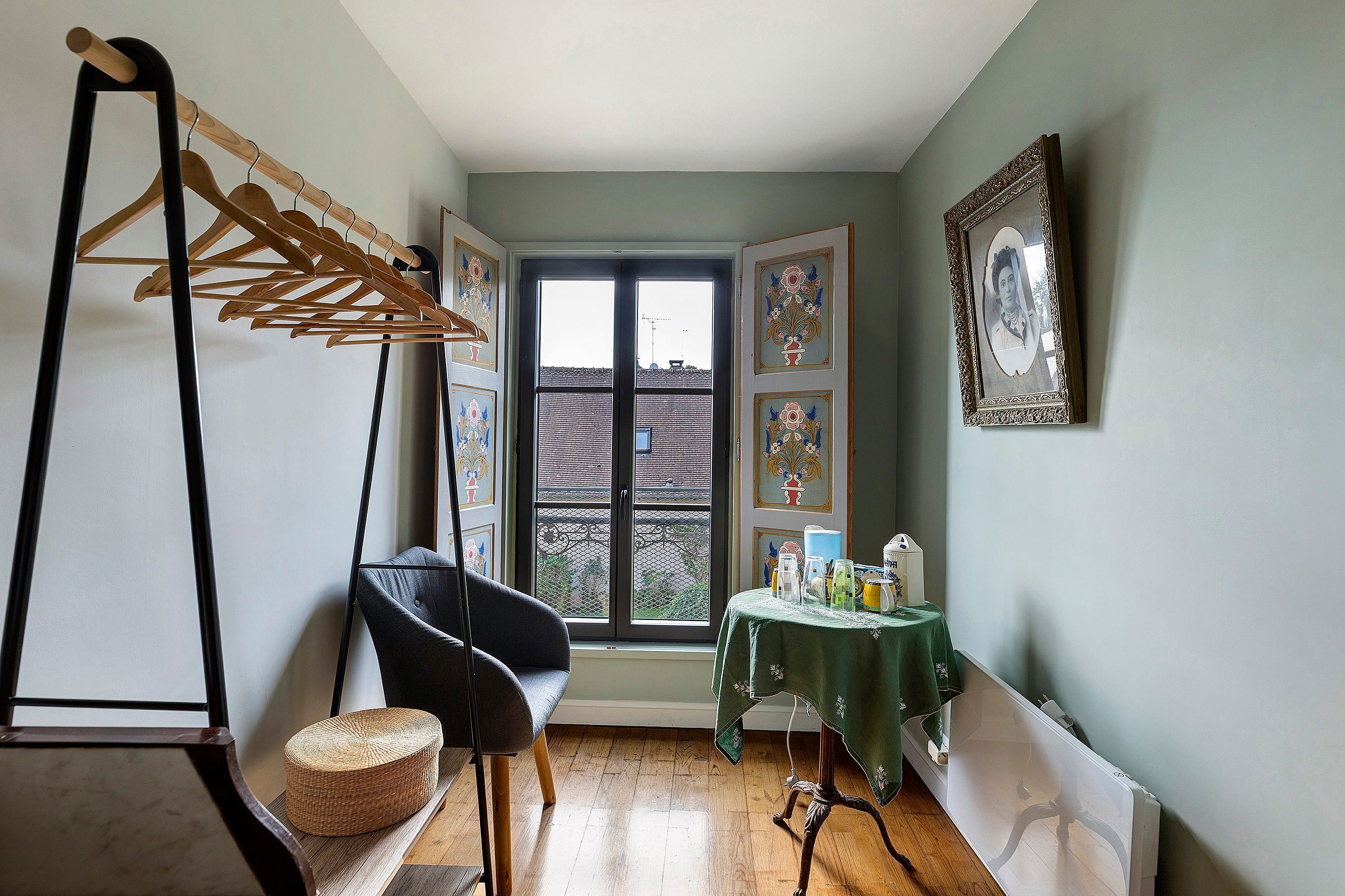 Een kamer met een houten droogrek, een zwarte stoel, een klein tafeltje met een groene tafelkleed en een ingelijst schilderij aan de muur.