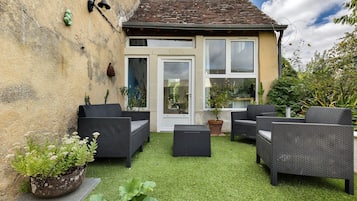 Une petite maison avec un toit en tuiles, une porte blanche et une fenêtre. On y trouve des meubles en osier et une plante en pot sur la pelouse.