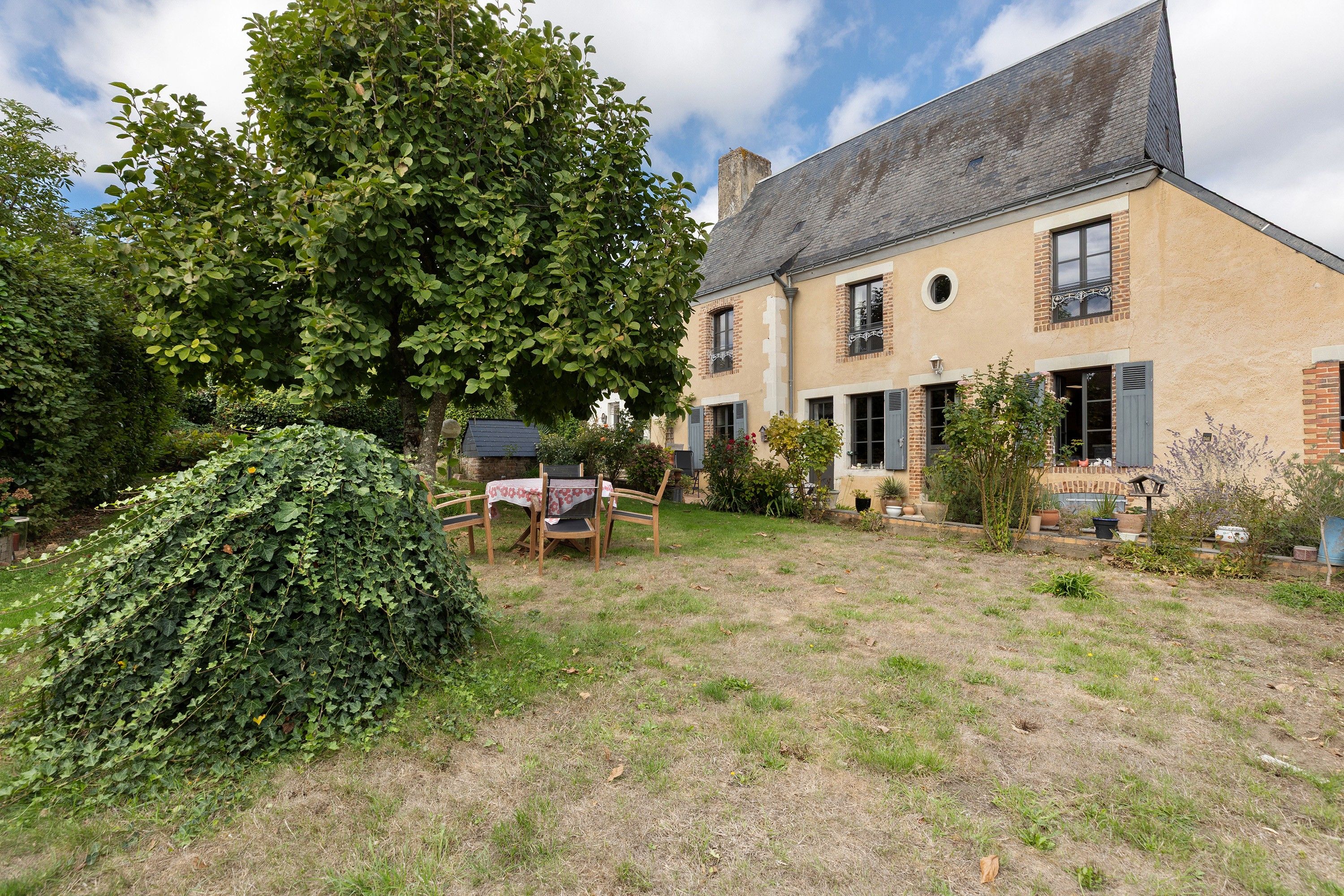 Une maison à deux étages avec une façade en briques, un grand arbre dans la cour et une table avec des chaises à l’extérieur.
