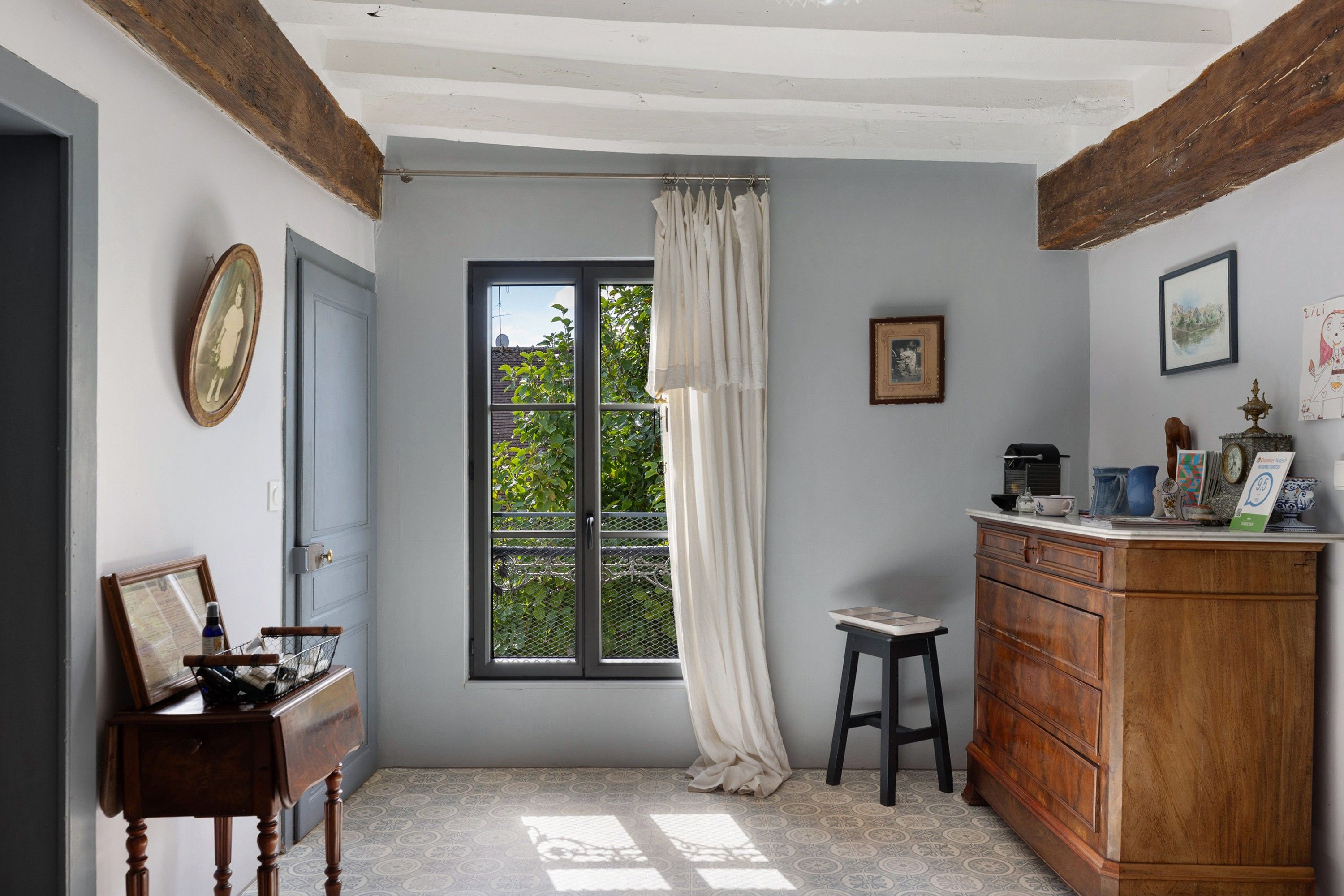 Une pièce avec une fenêtre, une porte, un petit tabouret, une armoire et un tableau encadré au mur.
