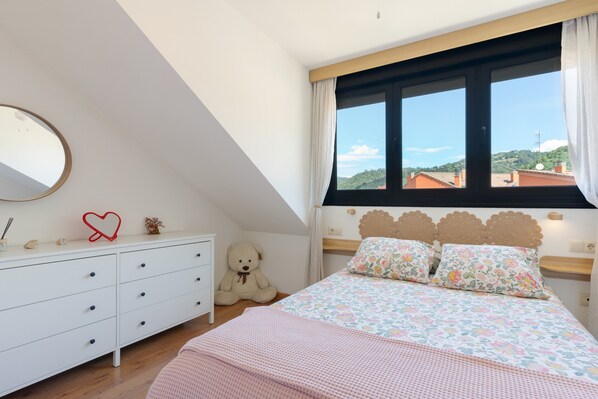 A bedroom with a large bed, a dresser, a teddy bear, a mirror, and a window with a view of the hills.