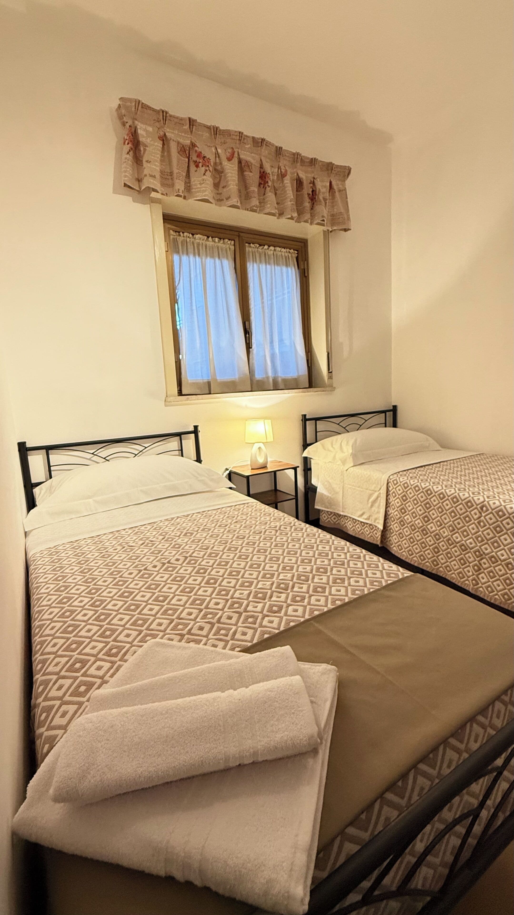 Quarto de hotel com duas camas, janela com cortinas, abajur e colcha estampada.