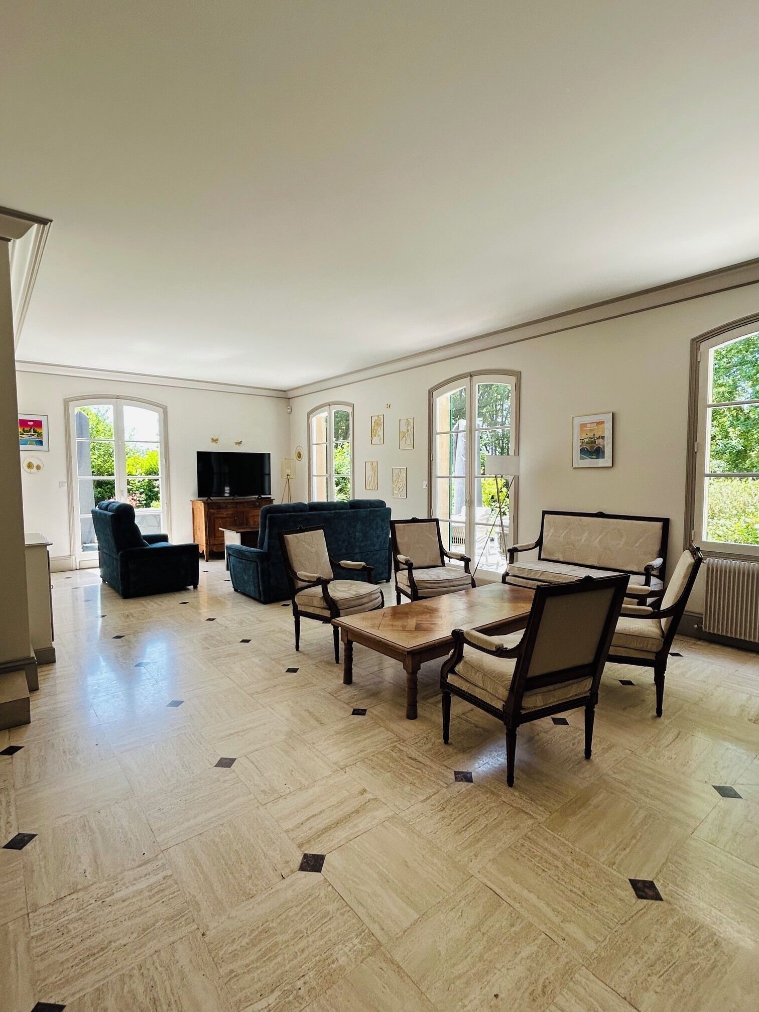 Un salon spacieux avec un grand canapé, une table basse et plusieurs fauteuils. On y trouve un téléviseur posé sur un meuble en bois, et la pièce est dotée de grandes fenêtres offrant une vue sur la verdure environnante.
