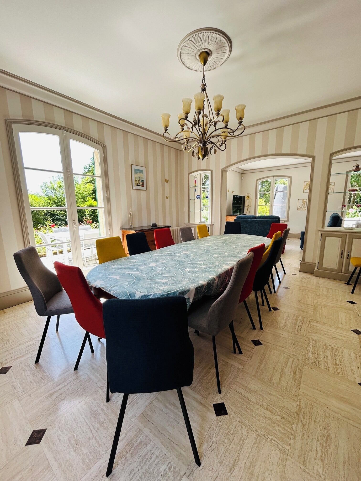 A dining area with a large table, chairs, a chandelier, and a view of the outdoors.