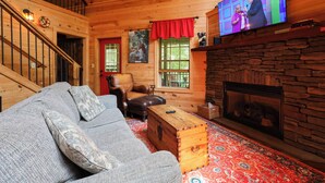 A cozy log cabin interior with a stone fireplace, a television mounted on the wall, a wooden staircase, and a comfortable seating area. - Firepit, hot tub, sleeps 7, exclusive hiking for resort, volleyball, peaceful! (Sevierville)