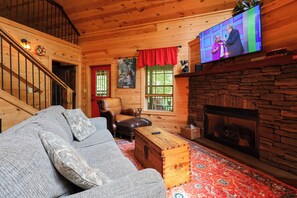 Ein gemütliches Blockhaus-Interieur mit Kamin aus Stein, einem an der Wand befestigten Fernseher, einer Holztreppe und einer komfortablen Sitzecke.
