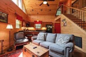 A cozy cabin interior with a wooden floor, a sofa, a red-framed window, a kitchen area, and a staircase. - Firepit, hot tub, sleeps 7, exclusive hiking for resort, volleyball, peaceful! (Sevierville)