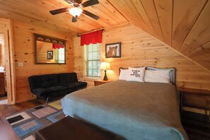 Ein gemütliches Schlafzimmer mit Holzbalkendecke, einem Bett mit grüner Bettdecke, einem schwarzen Sofa, einem Fenster mit Jalousien und einem Deckenventilator.