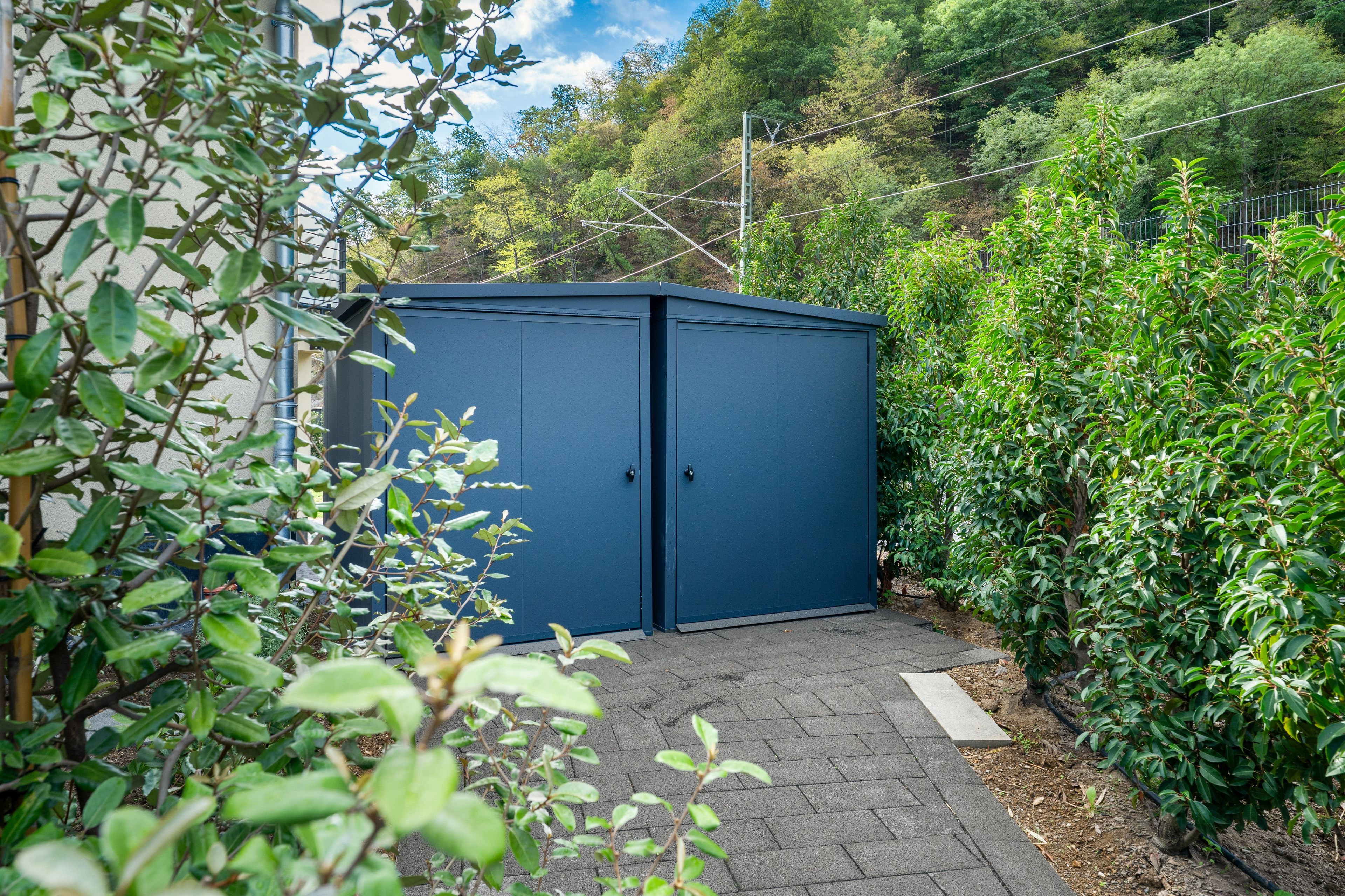 Een blauwe metalen schuur in een tuin, met een geplaveid pad er naartoe.
