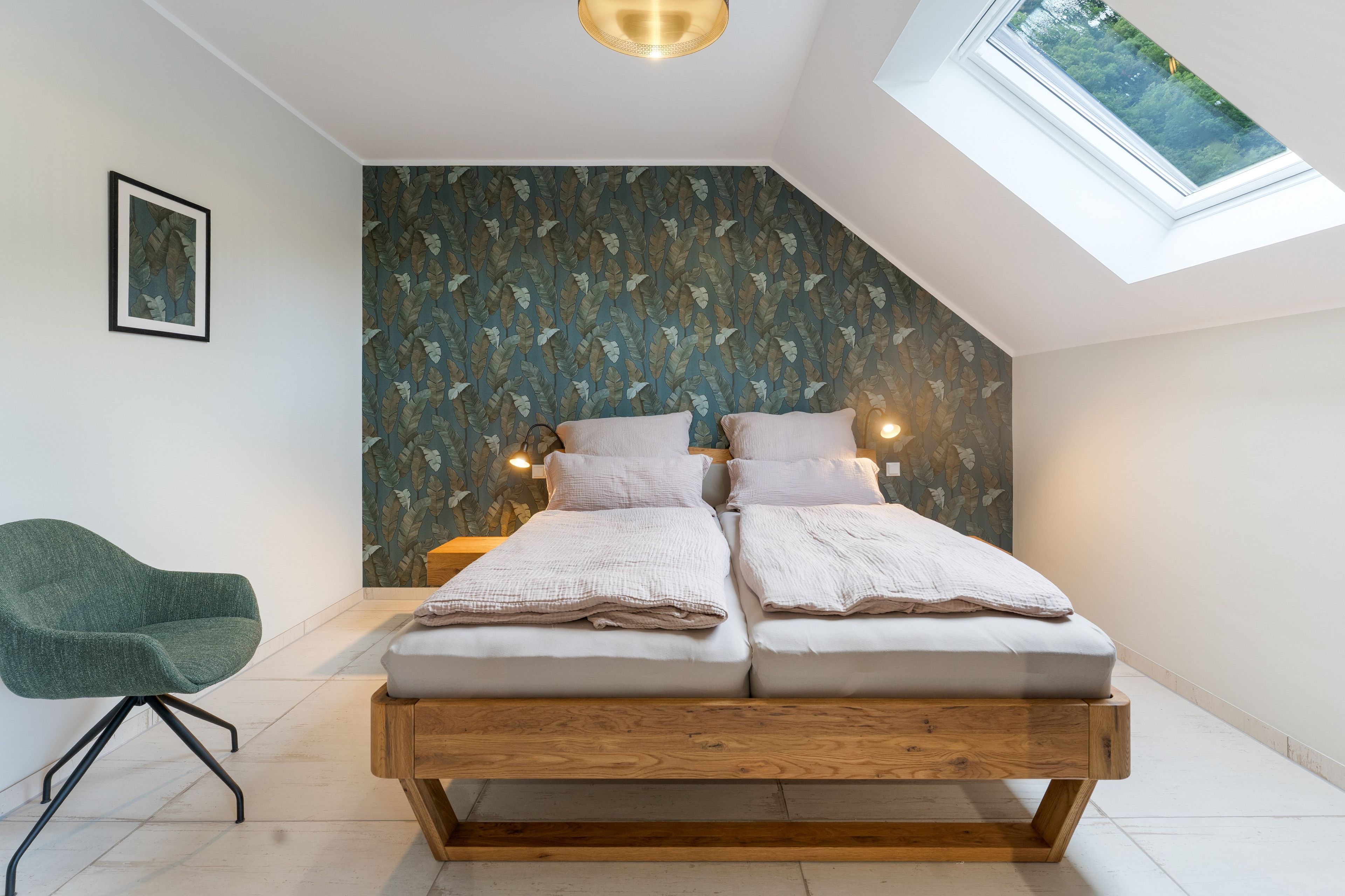 Ein Schlafzimmer mit Schrägdecke, einem Bett mit zwei Kissen, einem Sessel und einer Wand mit einem Blattmuster.