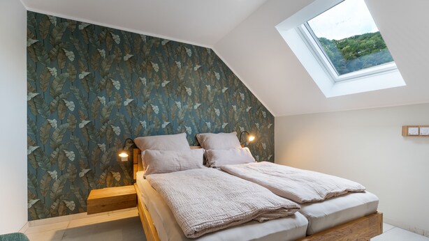 A bedroom with a sloped ceiling, a skylight, a bed with a wooden frame, and a wall with a leaf pattern.