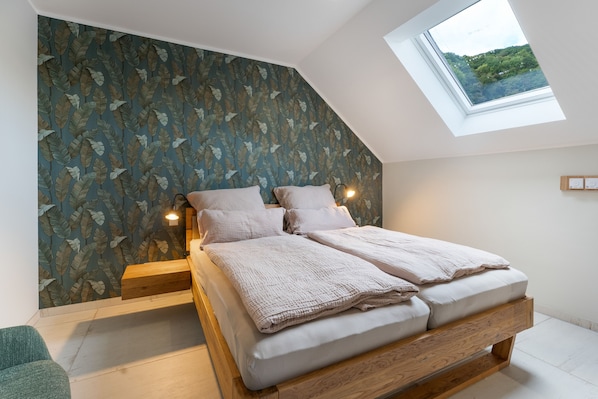 A bedroom with a sloped ceiling, a skylight, a bed with a wooden frame, and a wall with a leaf pattern.