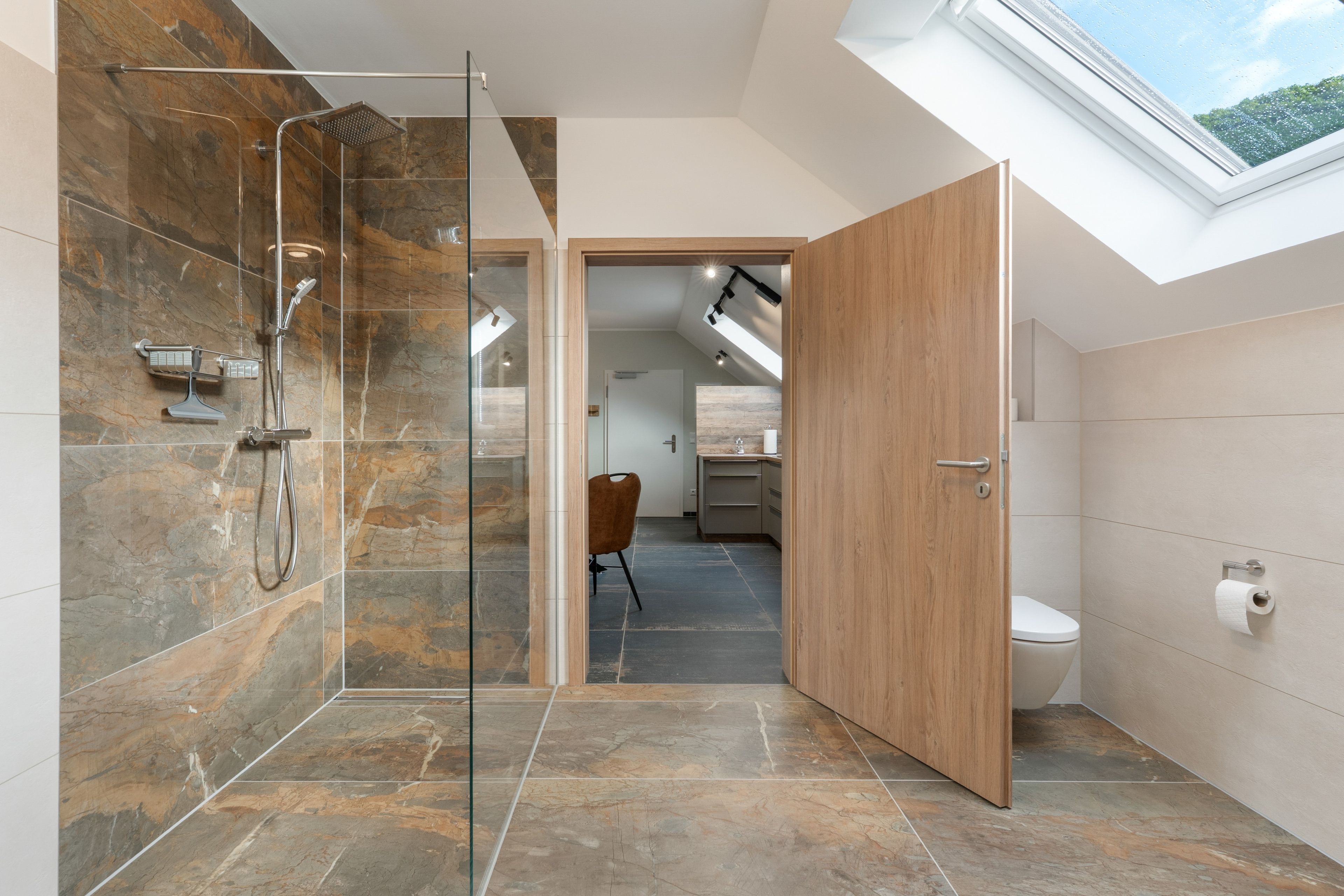 A modern bathroom with a glass shower enclosure, a wooden door, and a skylight.