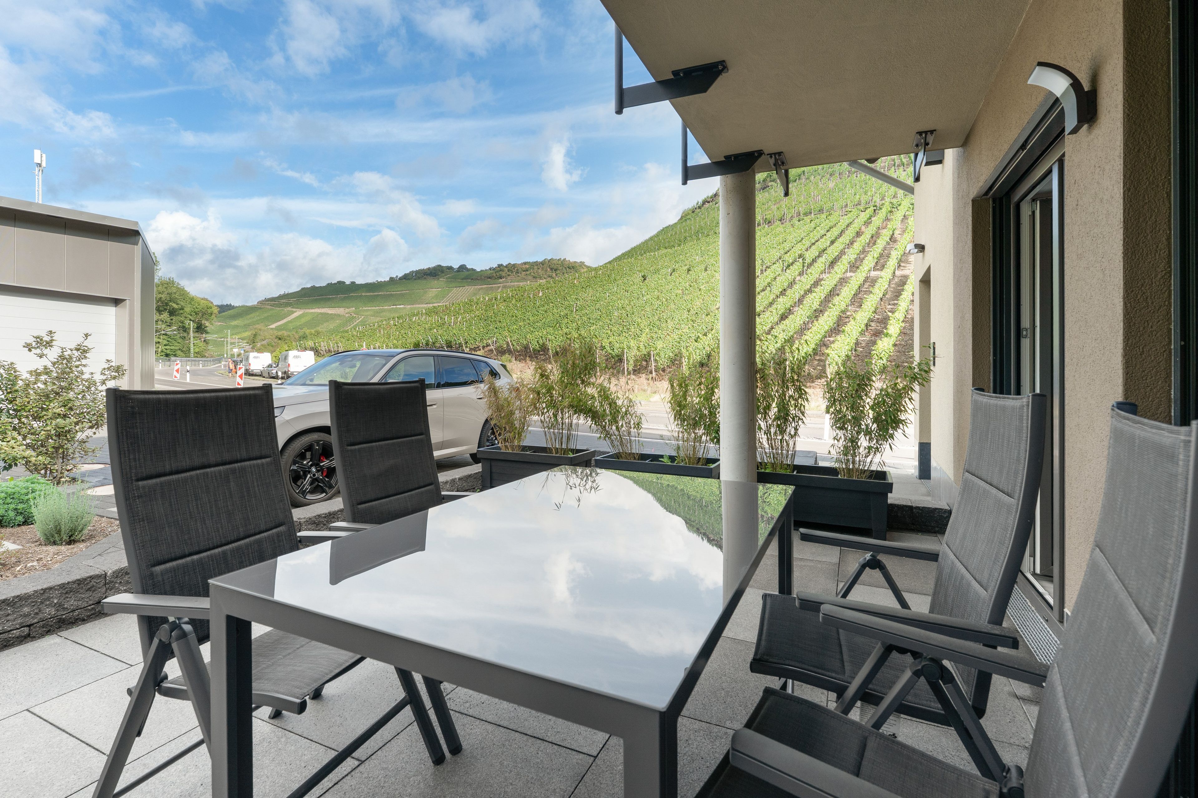 An outdoor patio with a table and chairs, a car parked nearby, and a view of a vineyard in the background.