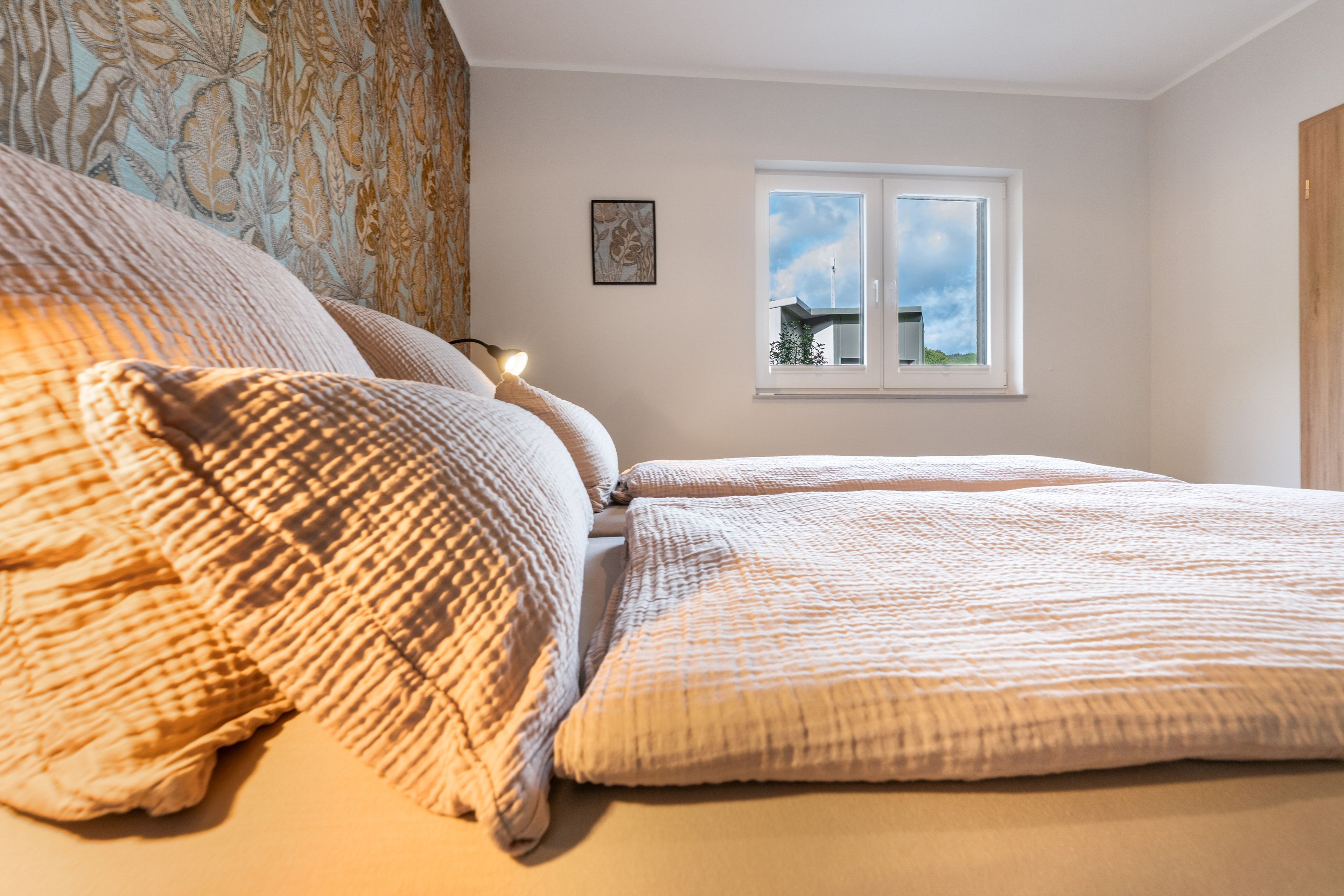 Een slaapkamer met een bed, een raam met uitzicht op een gebouw en een wand met een patroon gordijn.