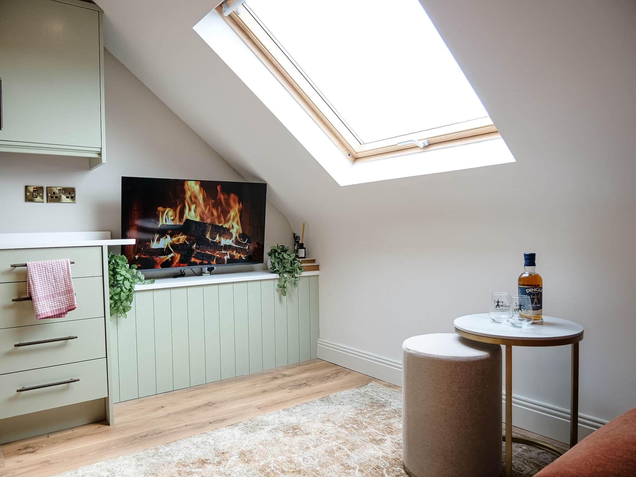 Une pièce avec un plafond en pente, une cheminée, une petite table ronde avec une bouteille et un verre, et une plante verte accrochée au mur.