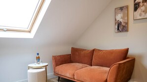 Une pièce chaleureuse avec un plafond en pente, un canapé marron, une petite table ronde avec des verres et un pouf blanc.