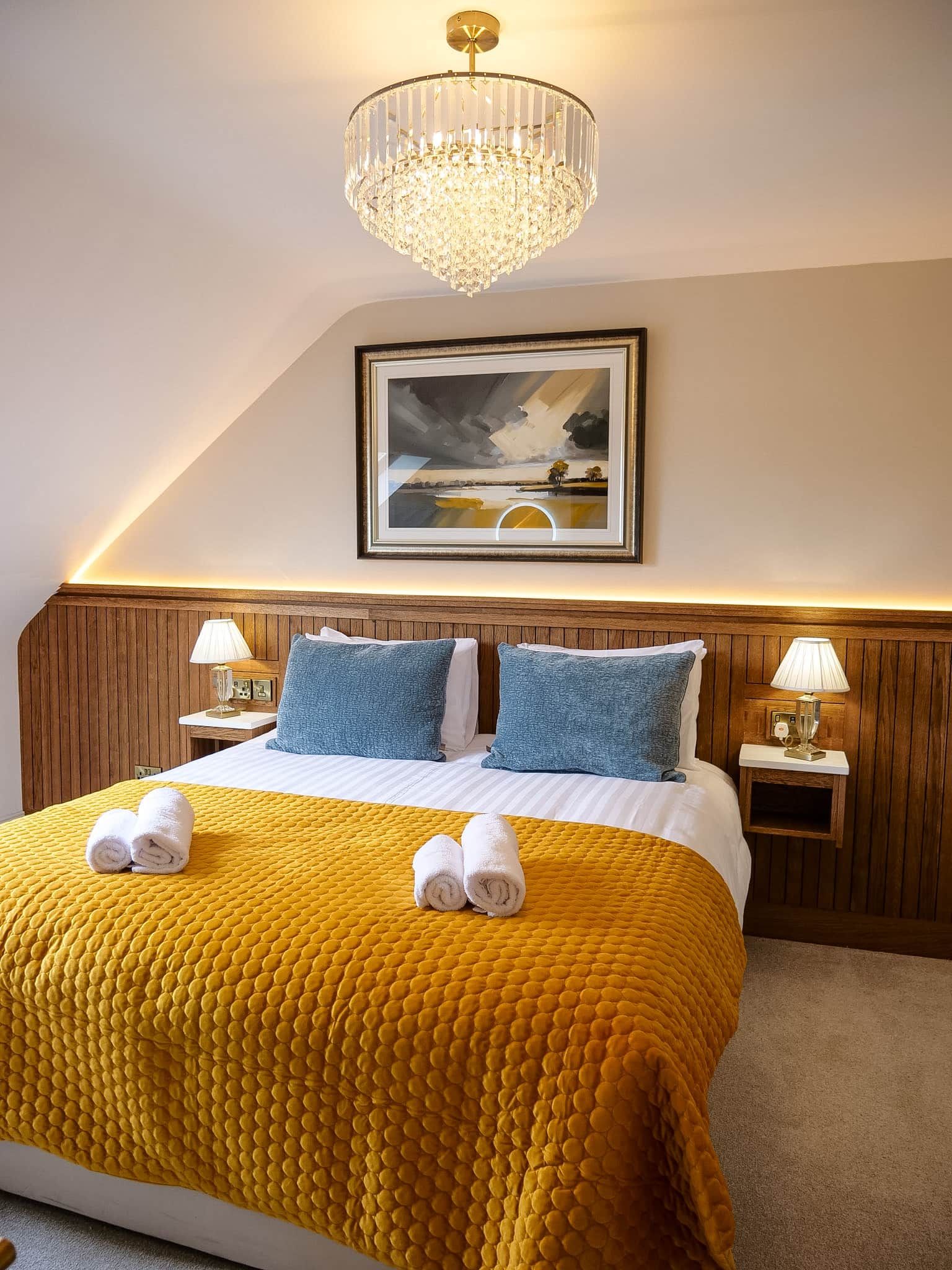 A hotel room with a large bed, two bedside lamps, a framed landscape painting, and a chandelier.