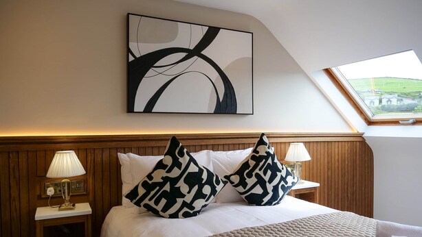 Une chambre d’hôtel avec un grand lit, deux lampes de chevet, un tableau abstrait moderne et une lucarne.