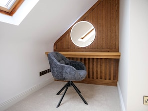 Una habitación con techo inclinado, pared de madera, espejo redondo, silla tapizada en gris y suelo alfombrado.