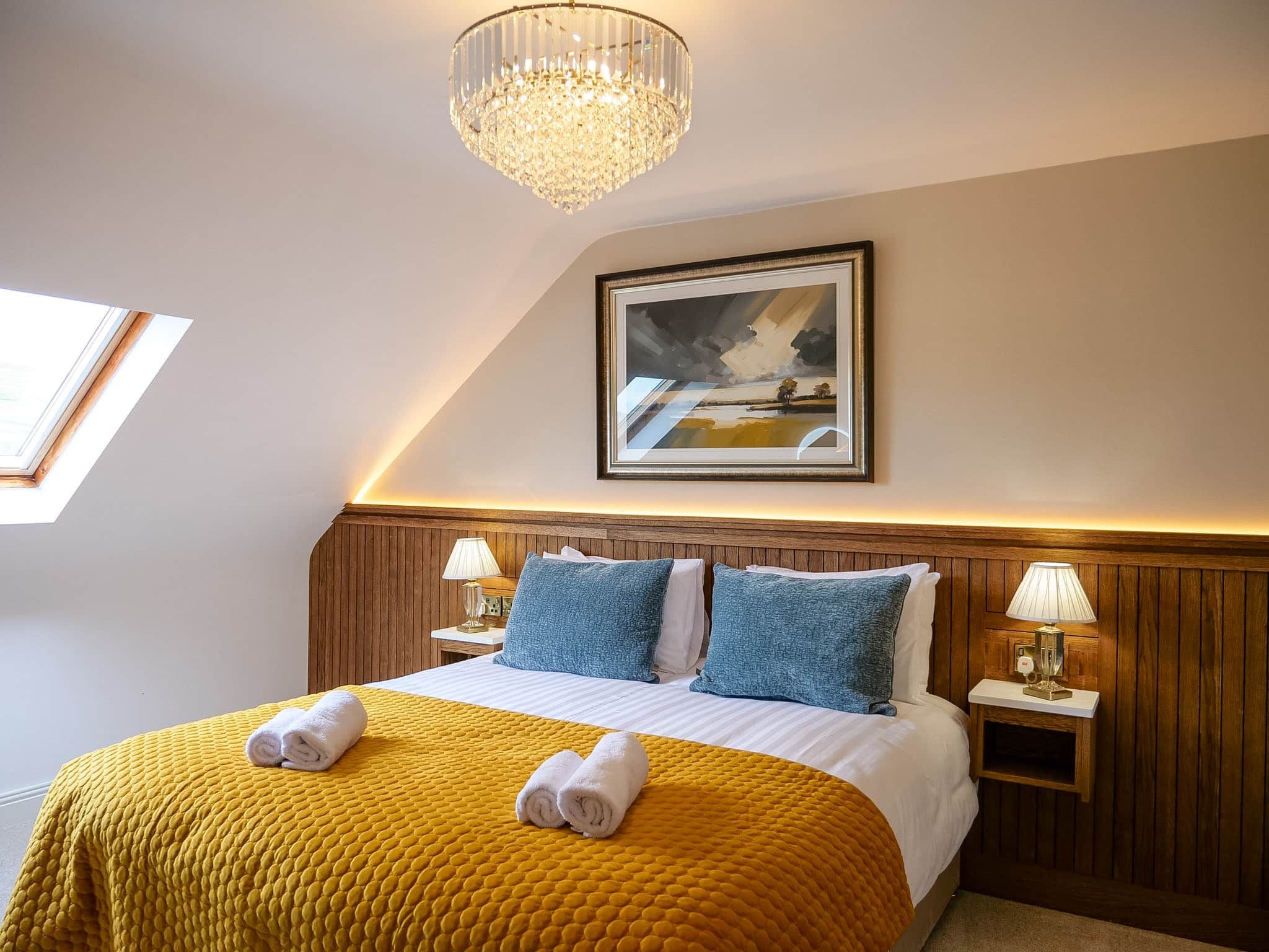 A hotel room with a bed, a painting, a chandelier, and a window.