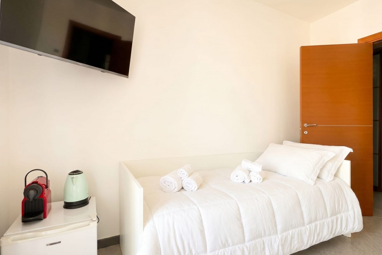 Ein Hotelzimmer mit Bett, einem an der Wand montierten Fernseher, einem kleinen Tisch mit Wasserkocher und einem roten Gerät sowie einer Holztür.
