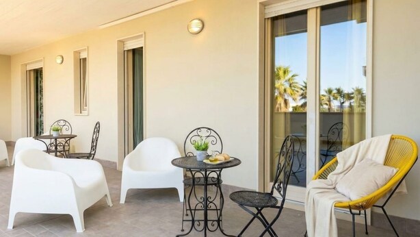 A covered outdoor seating area with white and yellow chairs, a small round table with a plant, and a glass door leading to a balcony with a view of palm trees.