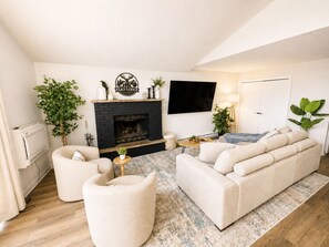 Ein Wohnzimmer mit Kamin, einer Sofagarnitur, einem an der Wand befestigten Fernseher und einem Blumentopf.