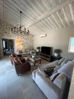 A spacious living room with a stone wall, a chandelier, a flat-screen TV, and a large sofa. - COTE D'AZUR - 4-BEDROOM, AIR-CONDITIONED STONE BASTIDE WITH POOL - SLEEPS 8 (Mougins)