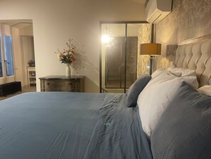 Une chambre avec un grand lit, une table de chevet ornée d’un vase de fleurs, une lampe et une vue sur l’espace de la douche.