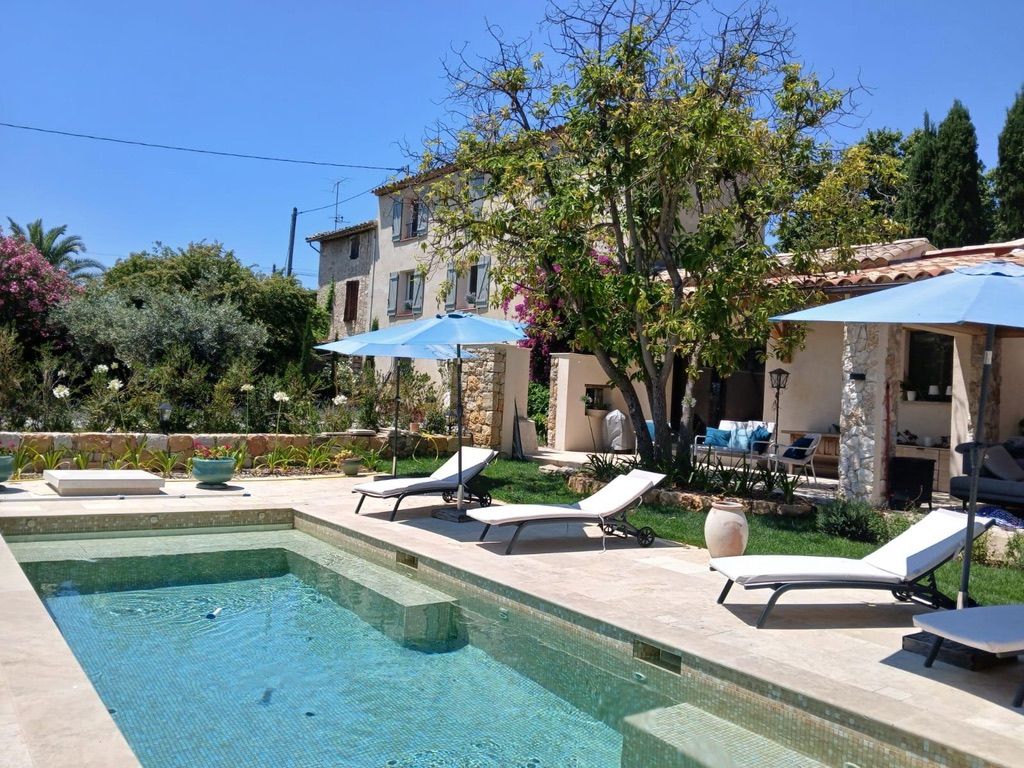 A pool area with lounge chairs and umbrellas, surrounded by a garden and a building with a tiled roof.