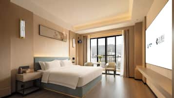 A hotel room with a bed, bedside table, a small table with chairs, a large window with a view, and a wall-mounted screen displaying hotel branding.