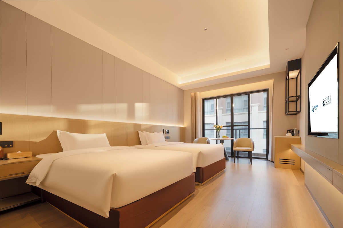 A hotel room with two beds, a desk, a chair, a small table with flowers, and a large window.