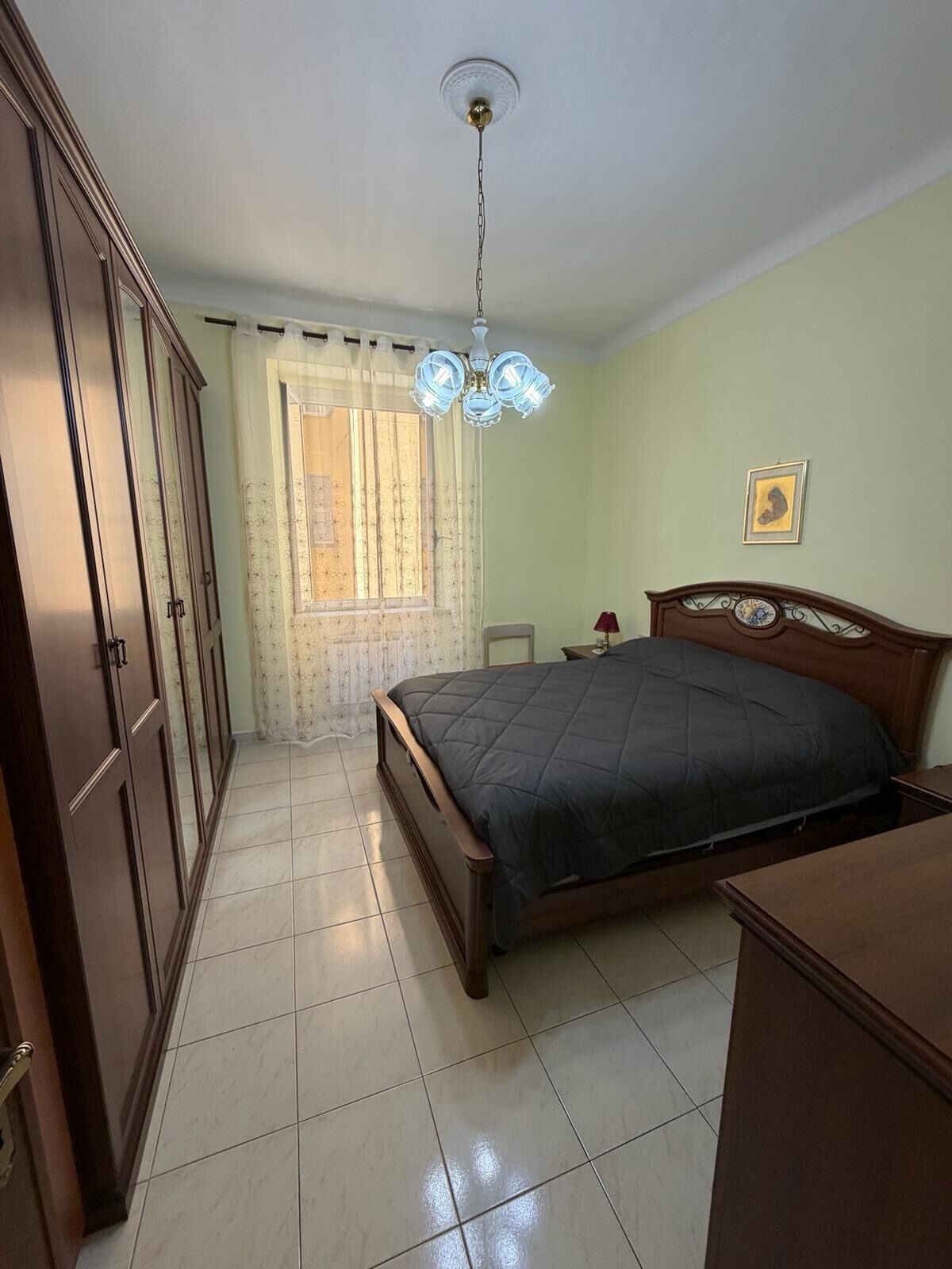 Une chambre à coucher avec un lit, une armoire, un lustre et une fenêtre avec des rideaux.