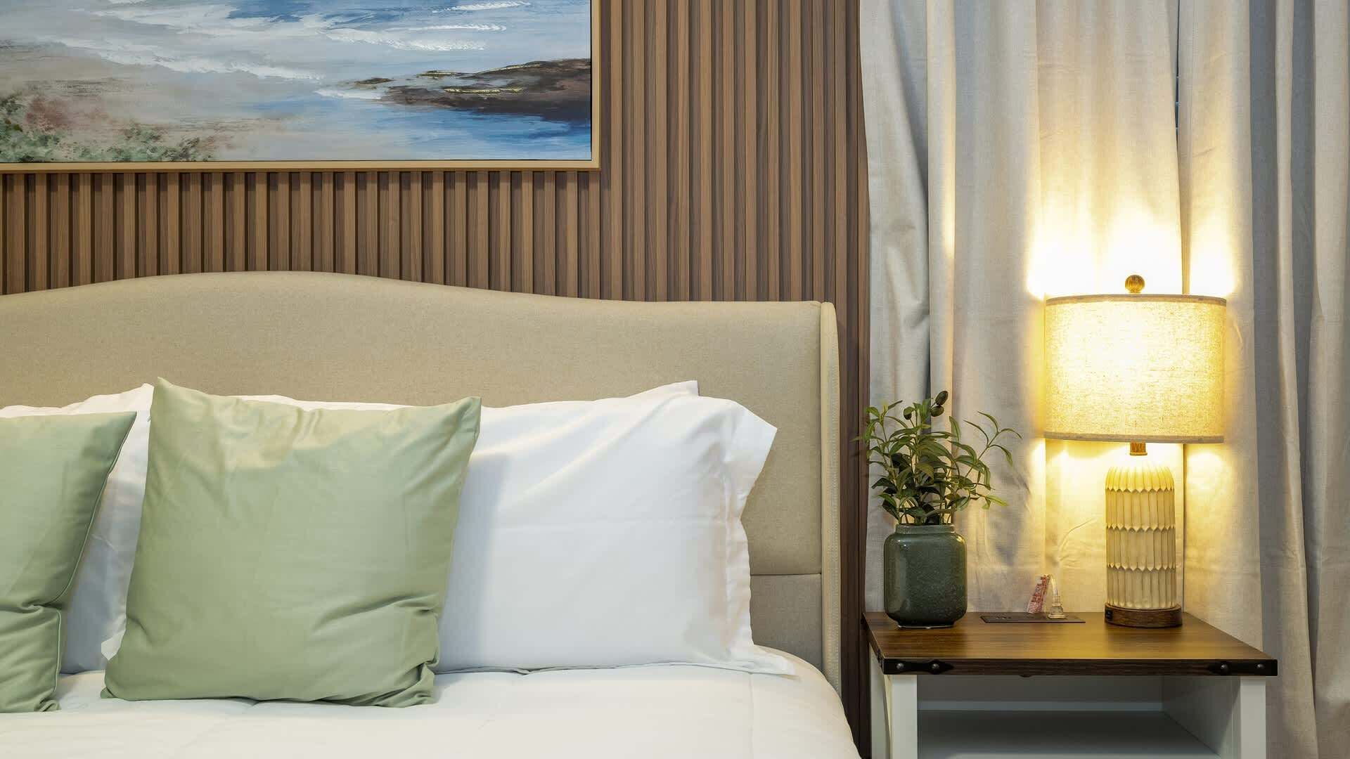 Ein Hotelzimmer mit einem Bett, Kissen, einem Nachttisch mit Lampe, einer Pflanze im Blumenvasen und einem Bild an der Wand.